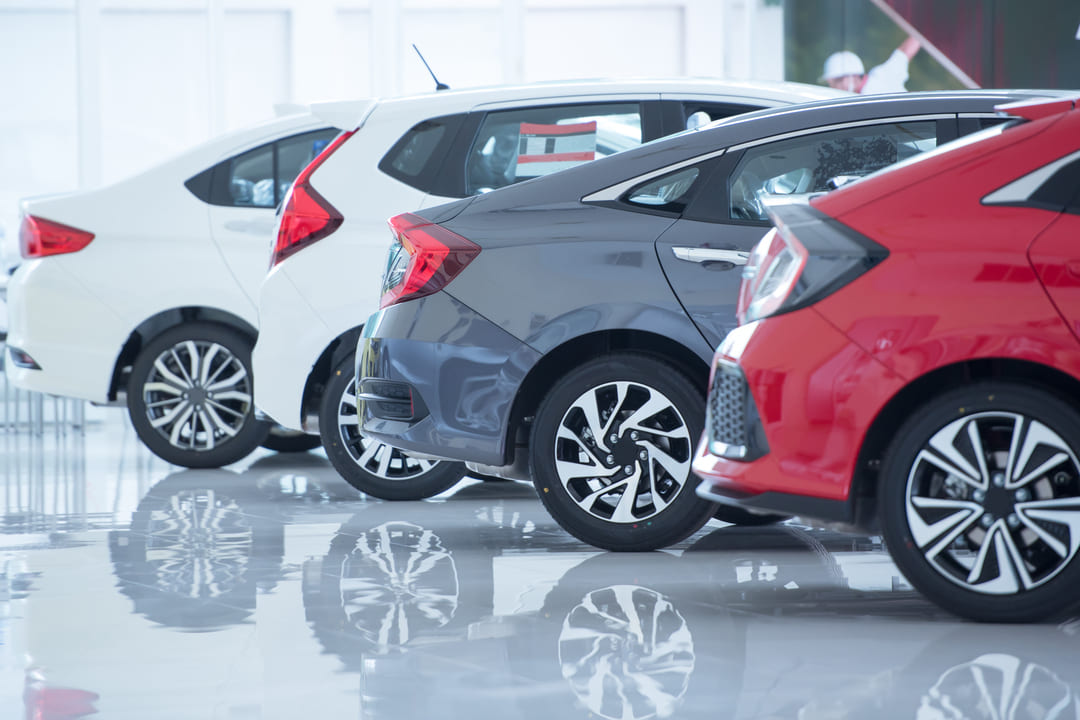 A imagem mostra uma fileira de carros novos expostos em um showroom automotivo. Os veículos são do mesmo modelo, mas de cores diferentes — branco, cinza e vermelho. Eles estão posicionados lado a lado, com destaque para a parte traseira e rodas esportivas, refletidas no piso brilhante do ambiente. O cenário é bem iluminado, com aparência limpa e sofisticada, típica de concessionárias.
