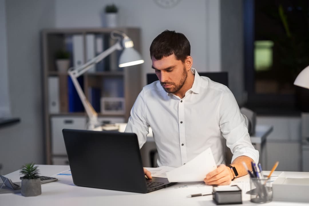 Homem analisando um laudo cautelar em frente ao notebook, em ambiente de escritório à noite.