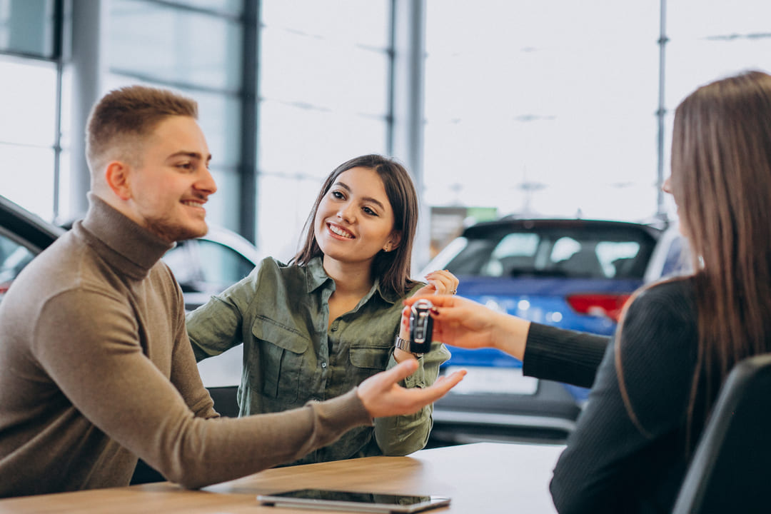 Jovem casal feliz recebendo chaves de carro de um vendedor em uma concessionária, simbolizando a compra de um veículo.