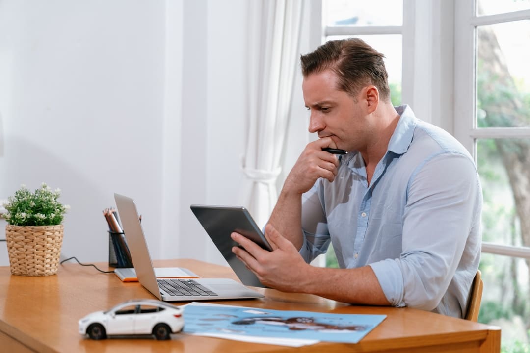 Homem analisando informações em um tablet, sentado à mesa com notebook, miniatura de carro e uma imagem impressa de automóvel, sugerindo consulta à tabela Fipe.
