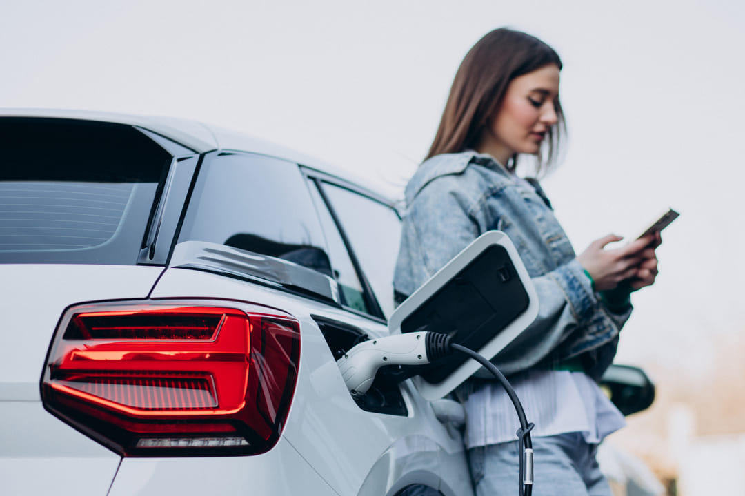 Mulher ao lado de um carro elétrico branco sendo carregado, enquanto usa o celular.