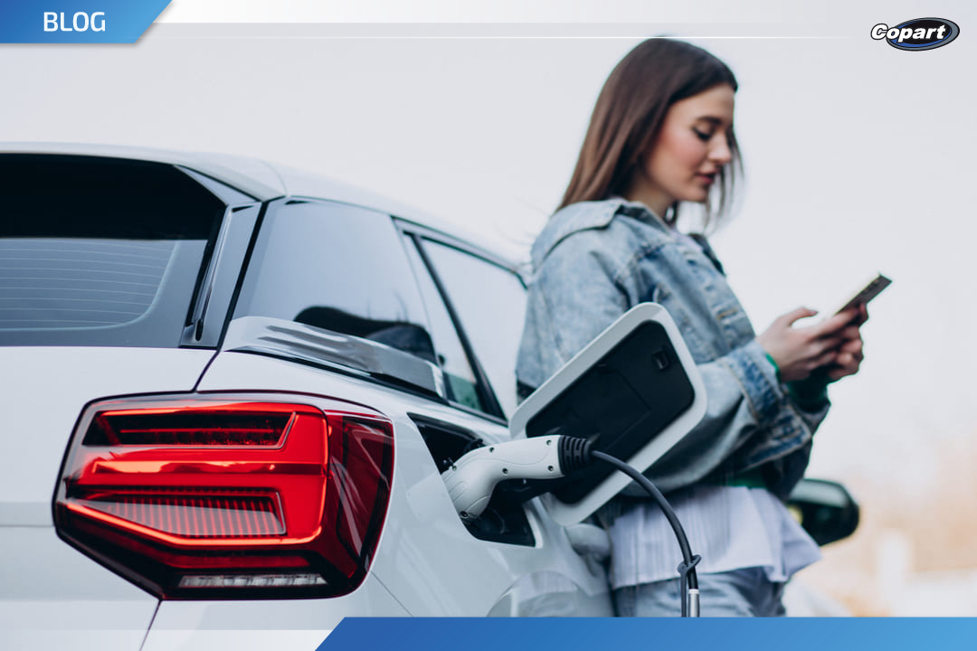 Mulher ao lado de um carro elétrico branco sendo carregado, enquanto usa o celular.