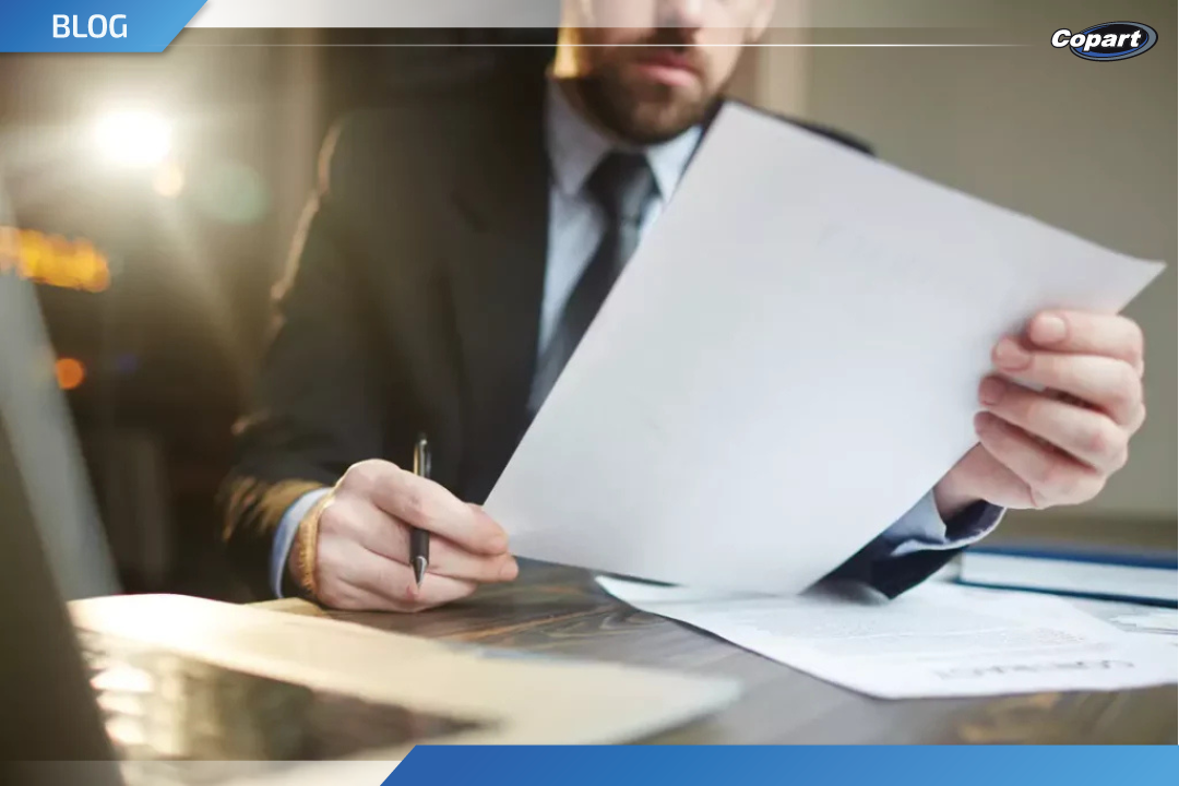 Homem de terno analisando um laudo cautelar sobre a mesa, com computador ao fundo.