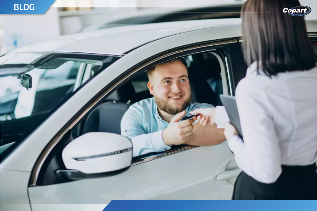 Homem sorridente dentro de um carro branco recebe a chave das mãos de uma vendedora, simbolizando a compra do primeiro carro.