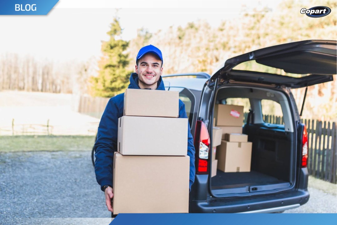 Entregador sorridente usando bon&eacute; azul e jaqueta carrega tr&ecirc;s caixas de papel&atilde;o, em frente a uma van com o porta-malas aberto e cheia de encomendas, em um ambiente externo.