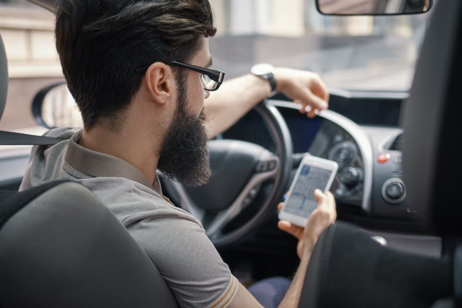 Homem com barba e óculos, sentado no banco do motorista de um carro, segurando um celular com aplicativo de navegação aberto, enquanto olha para frente com a mão esquerda apoiada no volante
