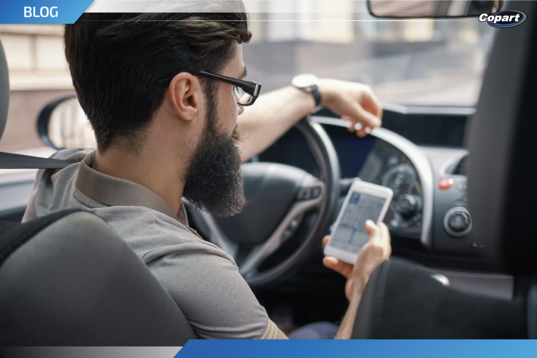 Homem com barba e óculos, sentado no banco do motorista de um carro, segurando um celular com aplicativo de navegação aberto, enquanto olha para frente com a mão esquerda apoiada no volante