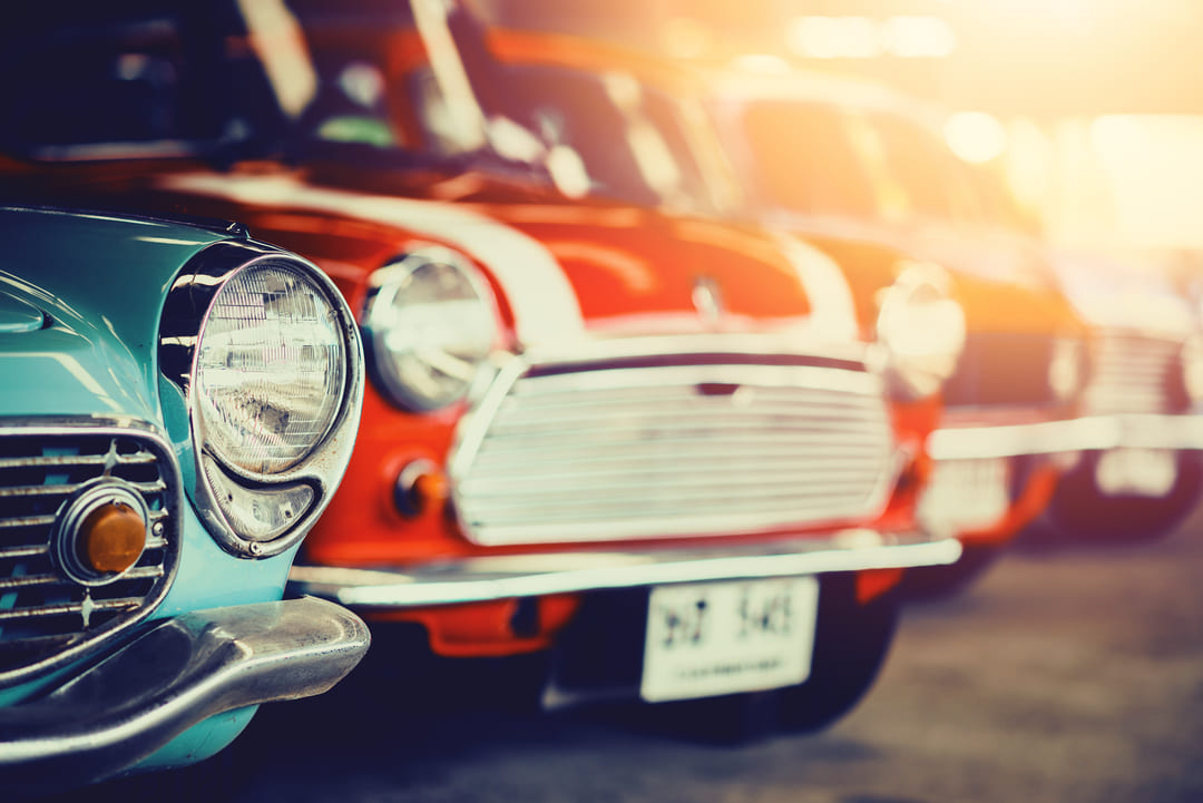 Fila de carros antigos coloridos estacionados, com destaque para os faróis e grades frontais sob luz suave do pôr do sol.