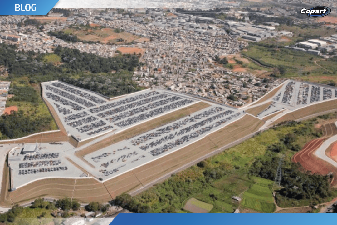 Vista aérea de um grande pátio de leilões de veículos, com centenas de carros estacionados em áreas delimitadas, cercado por áreas verdes e uma região urbana ao fundo.