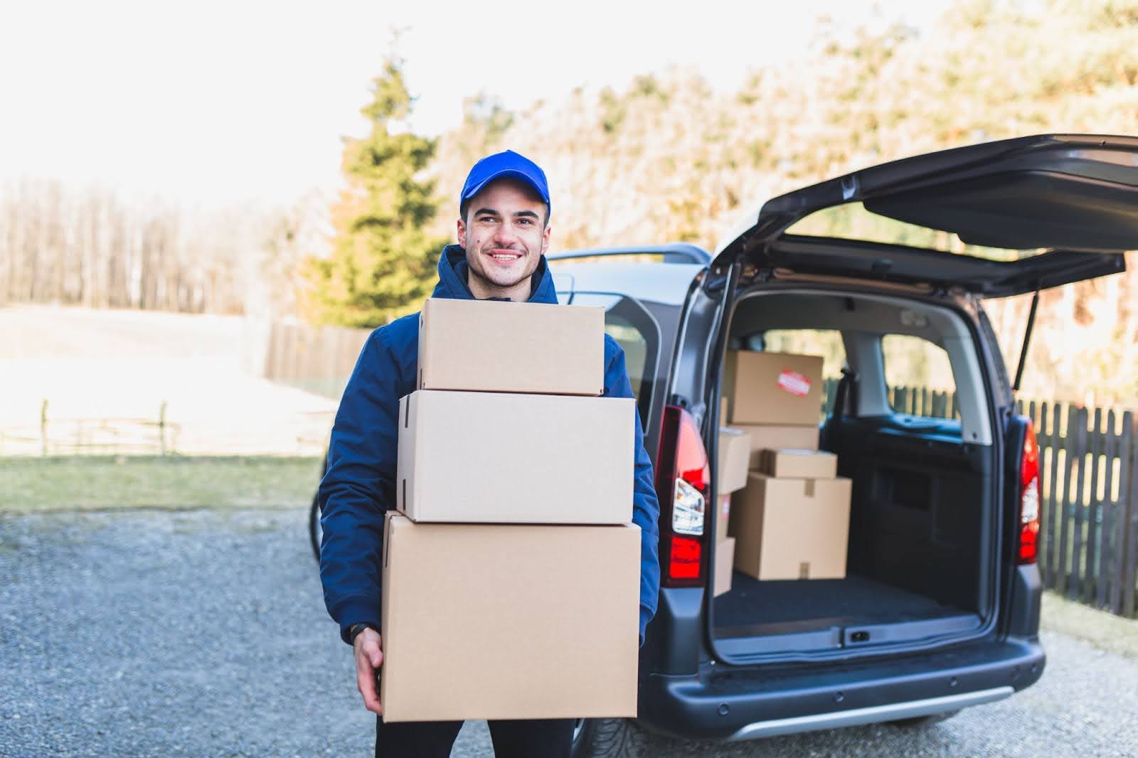 Entregador sorridente usando boné azul e jaqueta carrega três caixas de papelão, em frente a uma van com o porta-malas aberto e cheia de encomendas, em um ambiente externo.