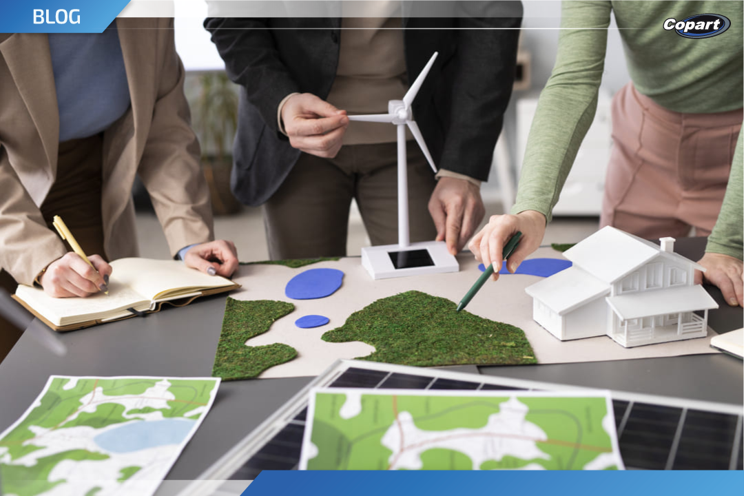 Tr&ecirc;s pessoas analisam uma maquete com elementos sustent&aacute;veis, incluindo miniatura de turbina e&oacute;lica, casa e &aacute;reas verdes, representando planejamento urbano e pr&aacute;ticas ESG.