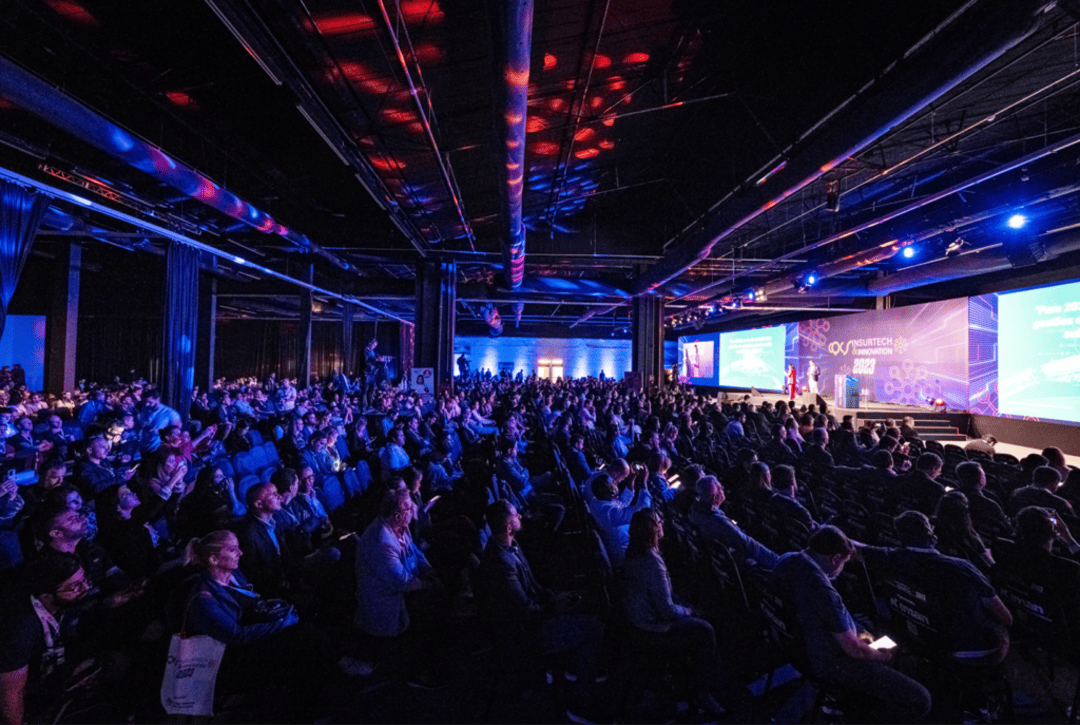 Público assistindo a uma palestra no CQCS Insurtech & Inovação 2025, em um auditório amplo e iluminado com luzes azuis e vermelhas, com telões exibindo apresentações no palco principal.