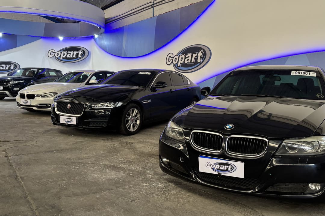 Fila de veículos premium, incluindo BMW e Jaguar, expostos em um galpão da Copart com iluminação azul, representando a Copart venda direta de veículos.