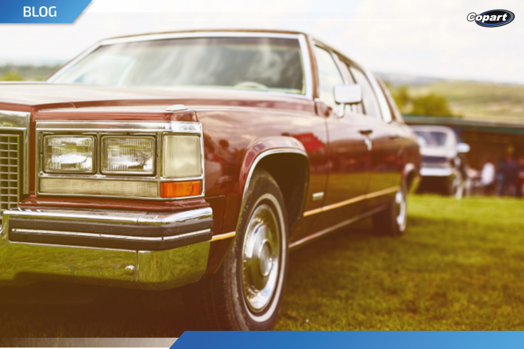 Automóvel clássico de cor vinho estacionado sobre gramado, visto em primeiro plano pela lateral dianteira, com outro veículo antigo desfocado ao fundo em um ambiente ao ar livre.