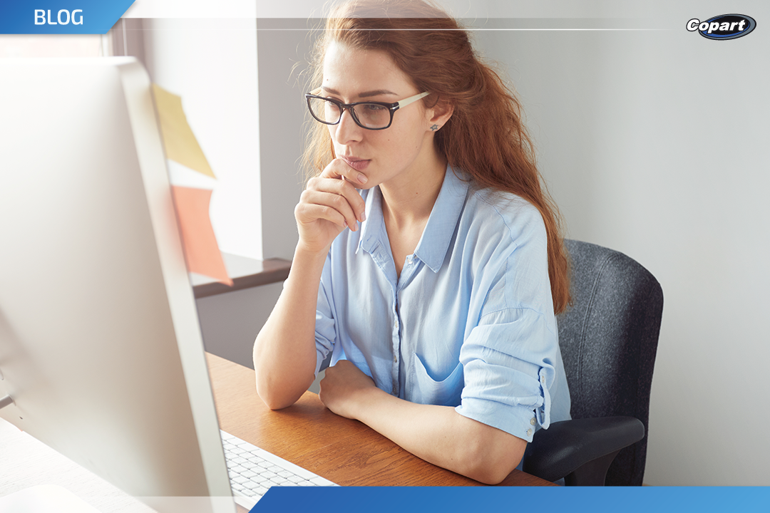 Mulher ruiva usando óculos, sentada em uma mesa de madeira, olhando atentamente para a tela do computador com a mão apoiada no queixo.